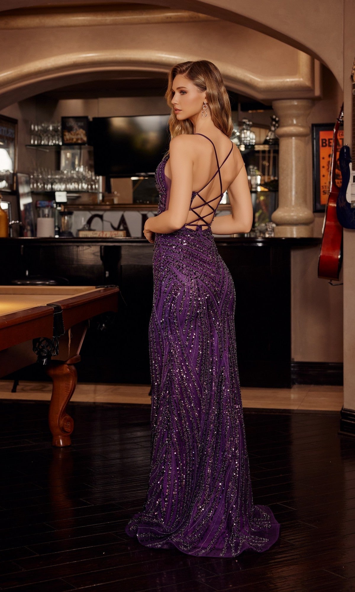A woman indoors wears the Nox Anabel C1836, a floor-length, sleeveless purple prom dress with sequin embellishments and a crisscross open back, as she faces away from the camera.